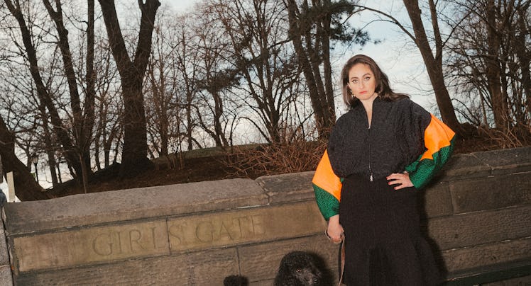 Catherine Cohen standing in front of Central Park in the fall with a black dog