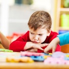 An upset child with head down on table as other children play with bright puzzle toys.