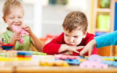 An upset child with head down on table as other children play with bright puzzle toys.