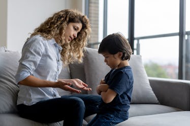 A mother sits on a couch and scolds her son, who crosses his arms and pouts.