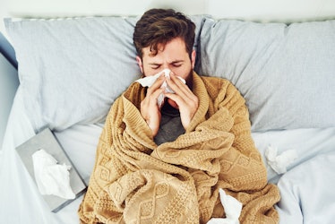 A sick man in a blanket on a couch blowing his nose with tissues.