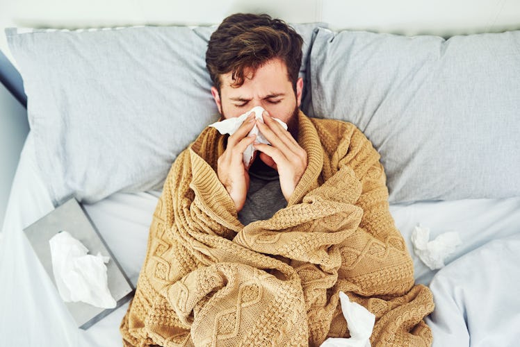 A sick man in a blanket on a couch blowing his nose with tissues.