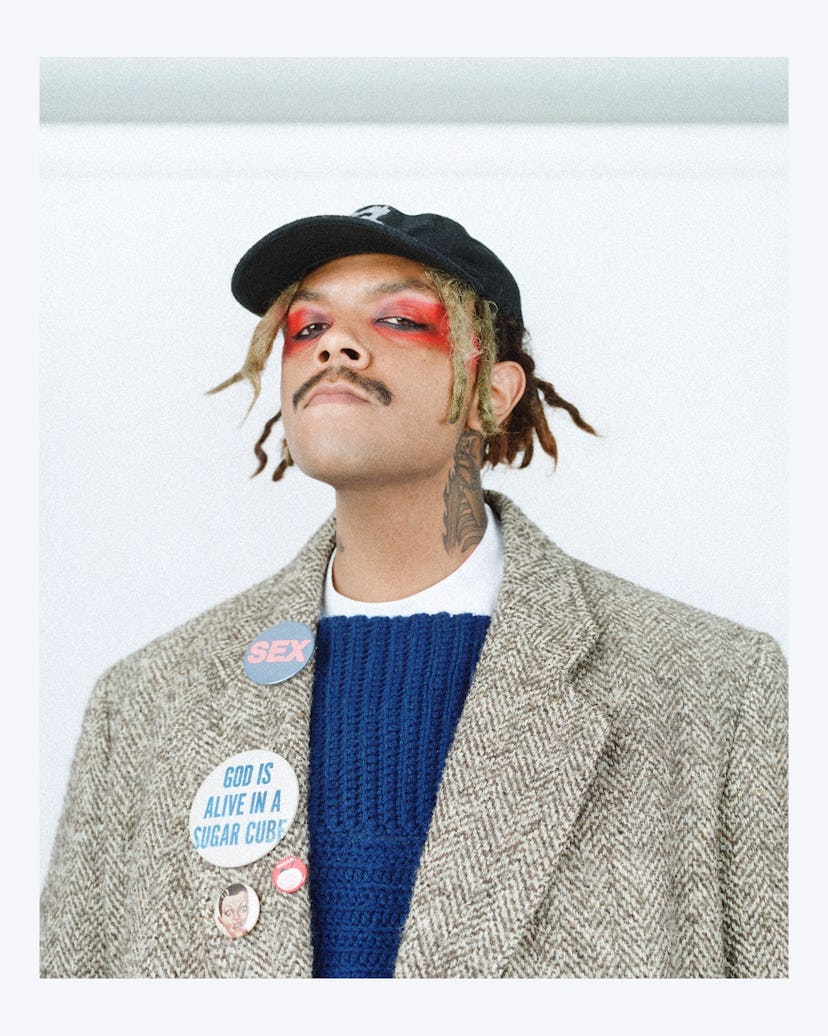 A portrait of Jean wearing red eye makeup, a black cap, a navy sweater, and tan blazer with buttons ...