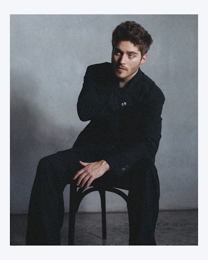A portrait of Froy wearing a black outfit, sitting on a chair