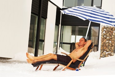 Man sunbathing outside in winter in chair under umbrella with snow on the ground.