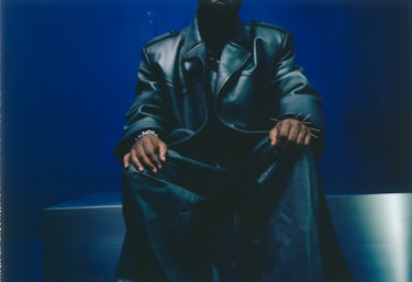 Kendrick Lamar sitting while wearing leather Prada coat, jumpsuit, Tiffany & Co. bracelets; his own ...