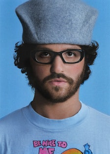 Eli Russell Linnetz wearing a blue t shirt, grey hat and black glasses and looking pensively directl...