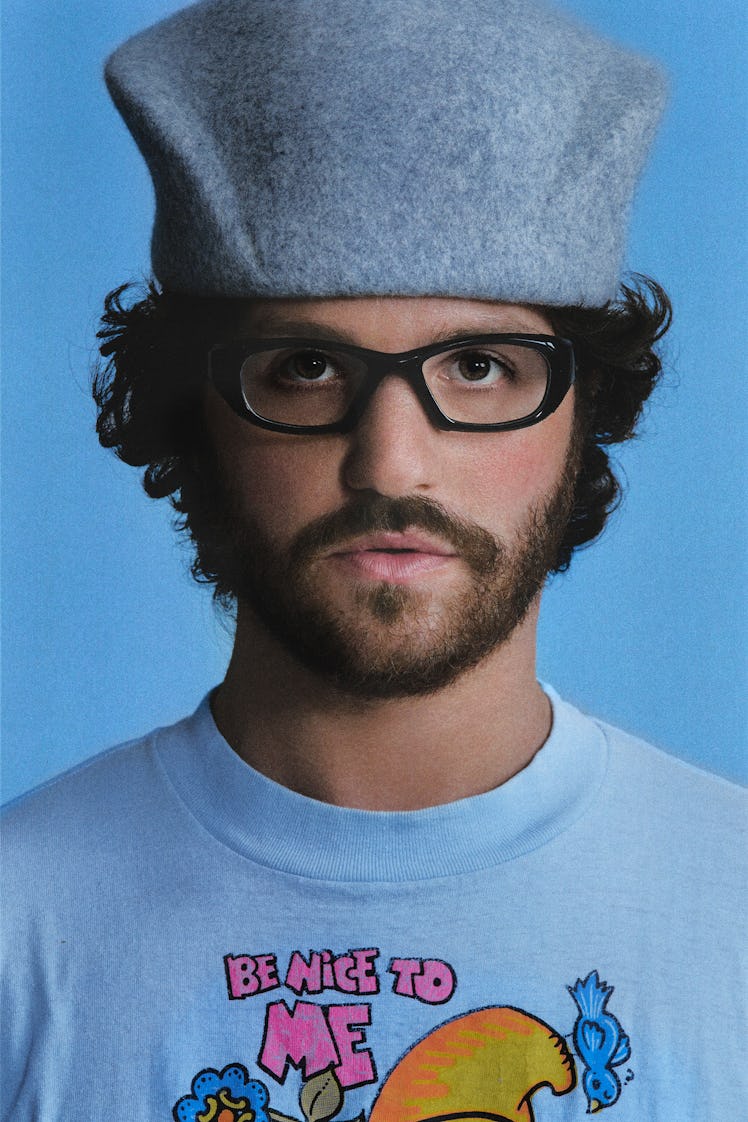 Eli Russell Linnetz wearing a blue t shirt, grey hat and black glasses and looking pensively directl...
