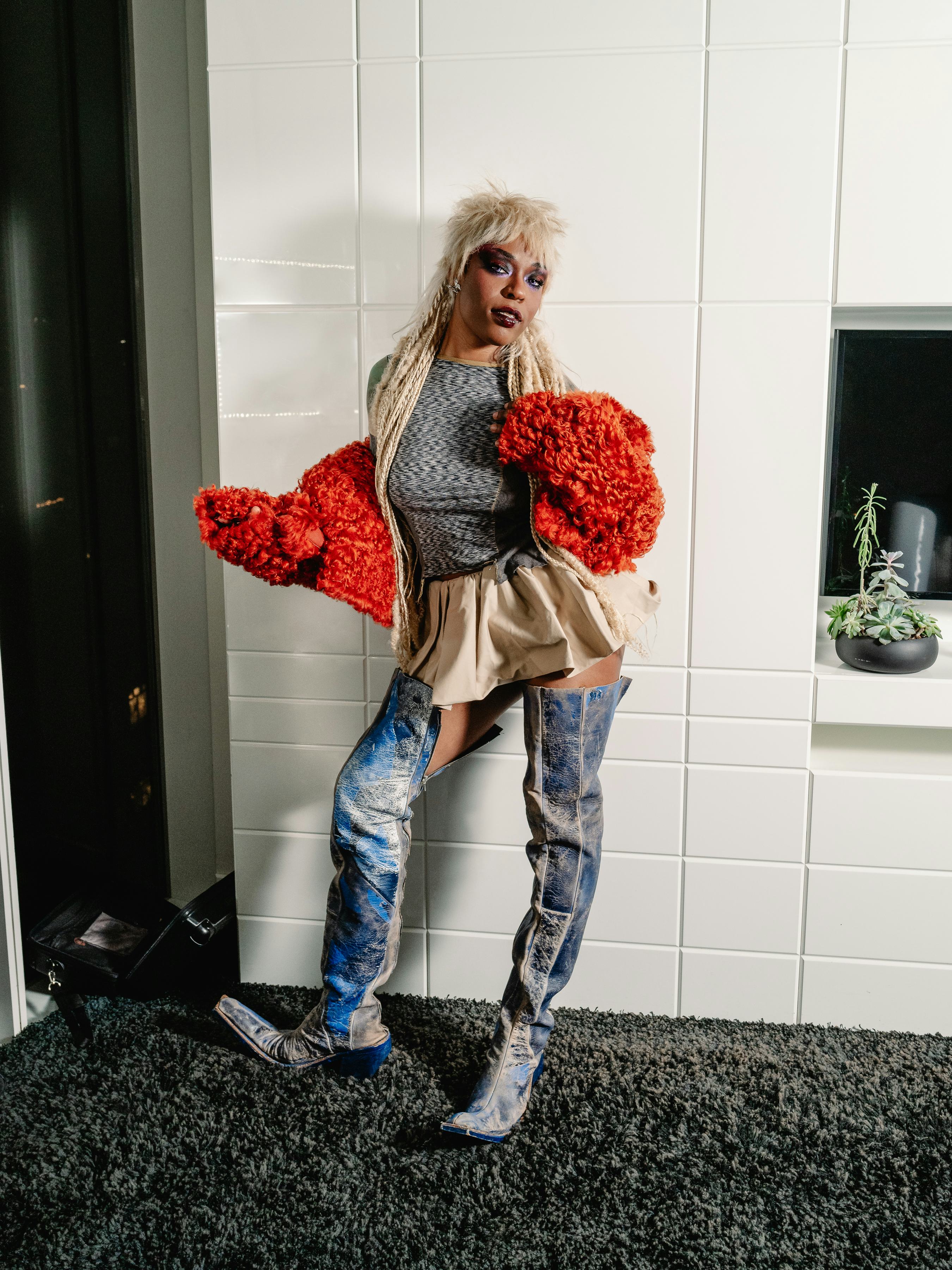 Sudan Archives wearing a red furry knit bolero, a shiny mini skirt, and thigh high boots