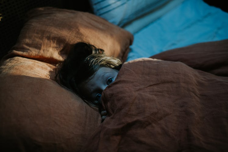 A girl hiding under her covers in bed in the dark, with scared eyes peeking out.
