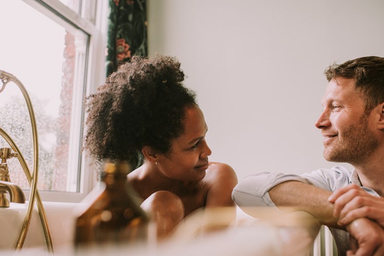 Woman in bathtub looks at her partner who is resting arm again tub