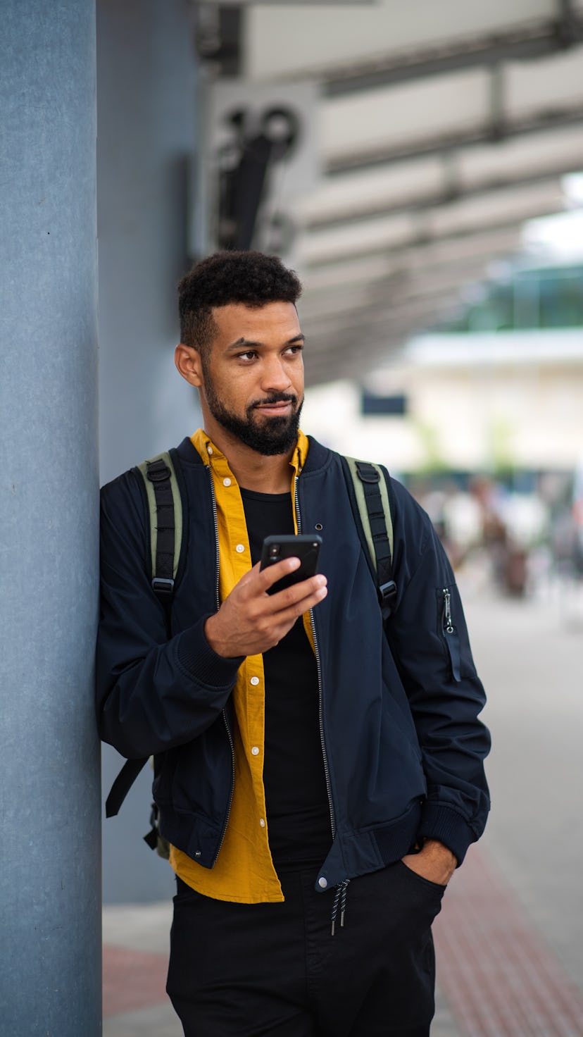 An attractive man leaning against a wall and holding his phone, one hand in his pocket.