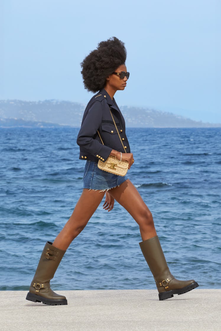 A woman walking in a black Celine blazer, denim mini skirt, and dark green boots