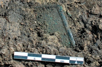 image of an artifact partially buried in soil with a ruler showing its comparative size, which is a ...