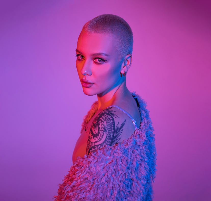 Model with a shoulder tattoo posing in front of a plain wall with purple lights.