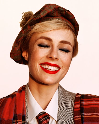 A model wearing a plaid blazer, necktie, and beret, smiling at the camera with a bold, red lip.