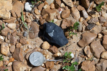 A black meteorite sits on the rocky ground next to a nickel for size. The meteorite is about 1.5 tim...