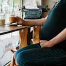 Midsection of a pregnant person sitting at a café window, drinking coffee.