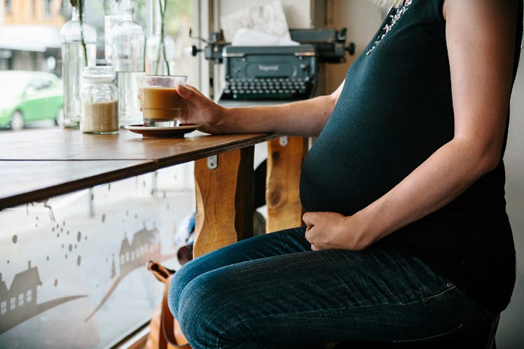 Midsection of a pregnant person sitting at a café window, drinking coffee.