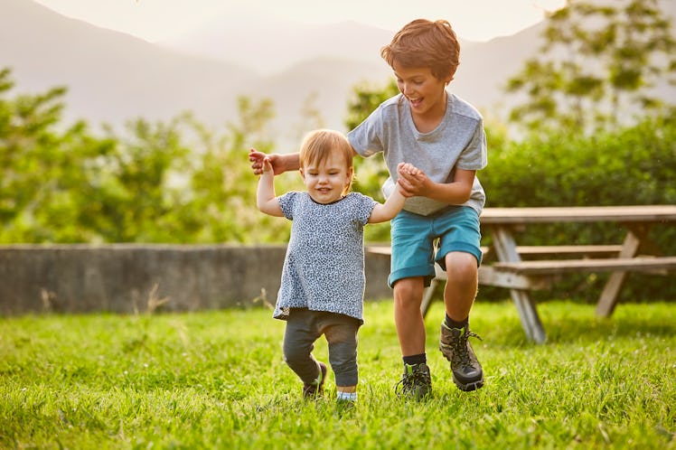 An older brother helping a toddler walk in the grass outside.