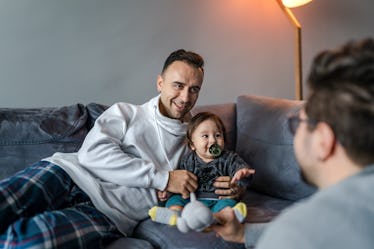 一位父亲坐在沙发上,带着蹒跚学步的孩子和孩子的另一位父亲交谈。