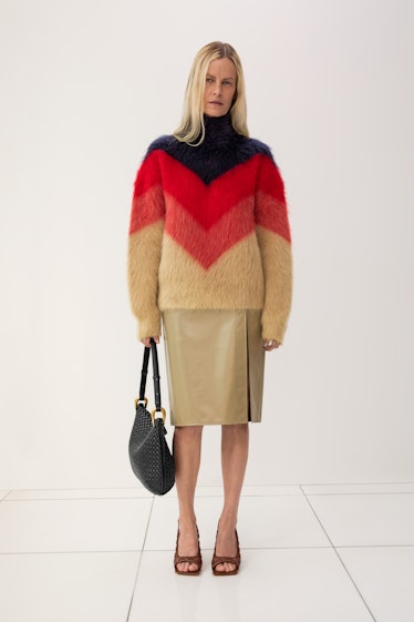 A blonde woman posing in a light brown skirt and brown, red, and black sweater by Bottega Veneta 