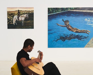 Antonio sitting on a yellow couch in his studio, playing guitar under two of his paintings