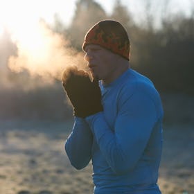 A man outside in cold weather, blowing visible breath into his hands, which are covered by mittens.