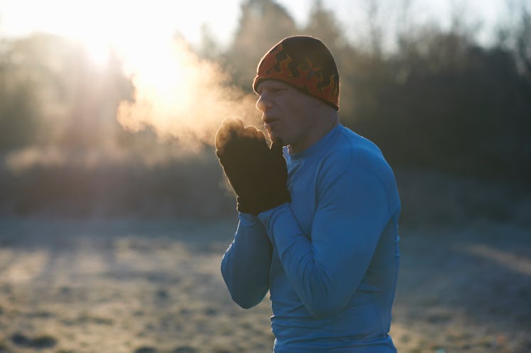 A man outside in cold weather, blowing visible breath into his hands, which are covered by mittens.