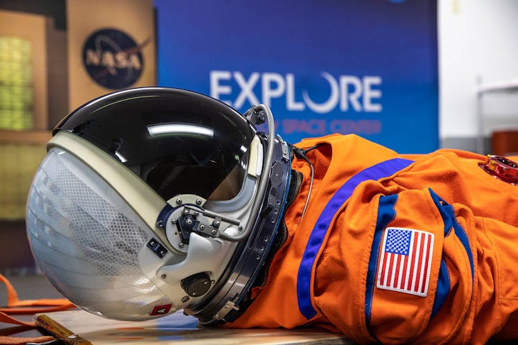 An orange spacesuit lies flat on a surface. It's visible from the upper torso on the right of the im...