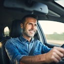 A man with a greying beard and dark hair smiling while driving his car.