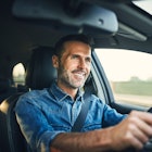 A man with a greying beard and dark hair smiling while driving his car.