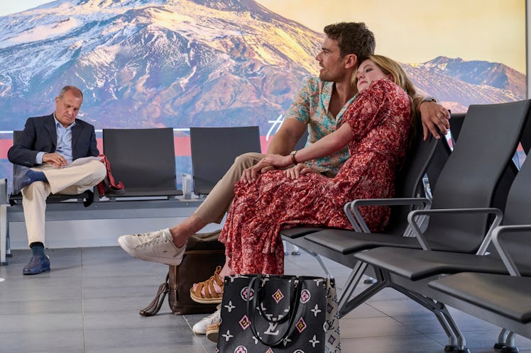 Theo James and Meghann Fahy sit in the airport with 'The White Lotus' volcano in the background of t...