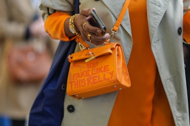 A guest wears a beige trench coat, an orange long sleeves long dress, gold rings, a gold watch, an o...