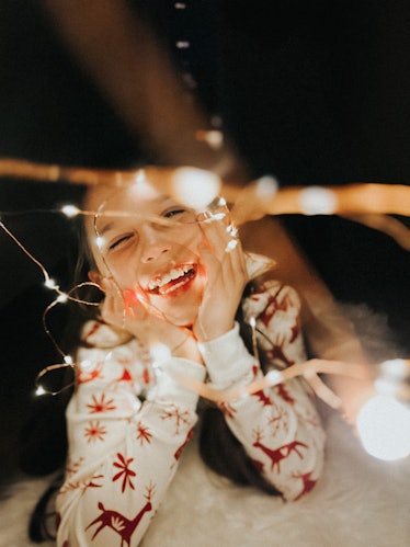 A young girl in Christmas pajamas lays on the floor, chin on her hands, smiling, with her face frame...