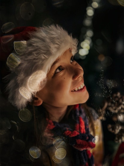 A young girl in a Santa hat smiles up at Christmas lights out of frame, with bokeh light spots all a...