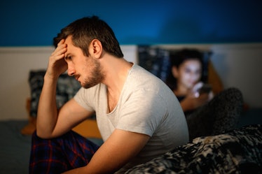 An annoyed man sits on a bed his his pajamas while his wife is on her phone.