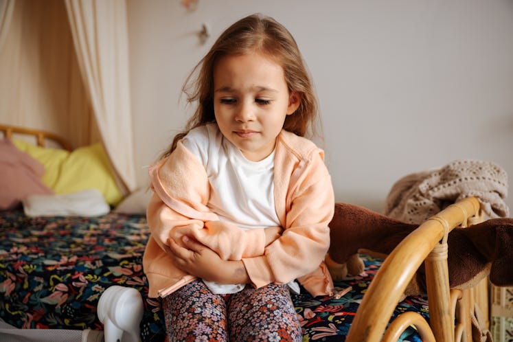 A child with a stomachache sitting on a bed, clutching her stomach.