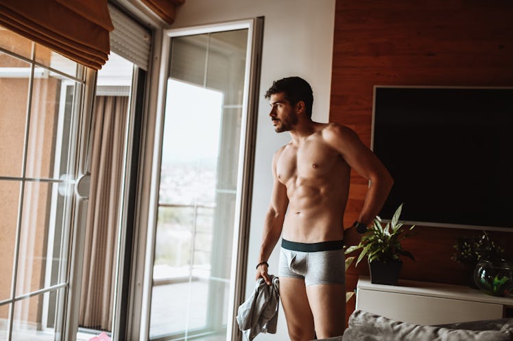 A man in his bedroom wears only underwear, holding his pants in his hand.