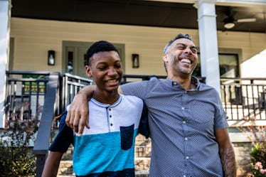 A dad has his arm around his son's shoulder while they laugh outside their home.