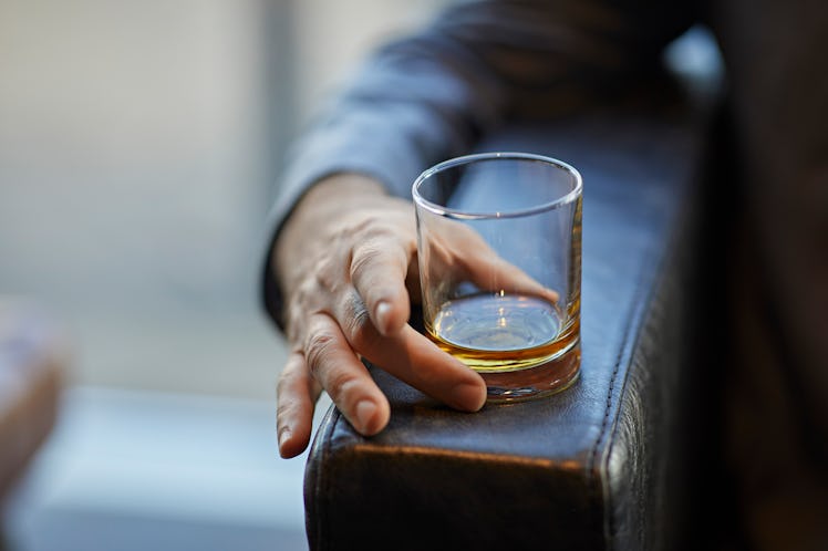 Close up of a glass of whiskey resting on the end of the arm of a leather chair as a man holds it.