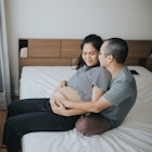 A man sits behind his pregnant wife on their bed, holding her.