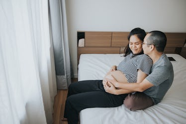 A man sits behind his pregnant wife on their bed, holding her.