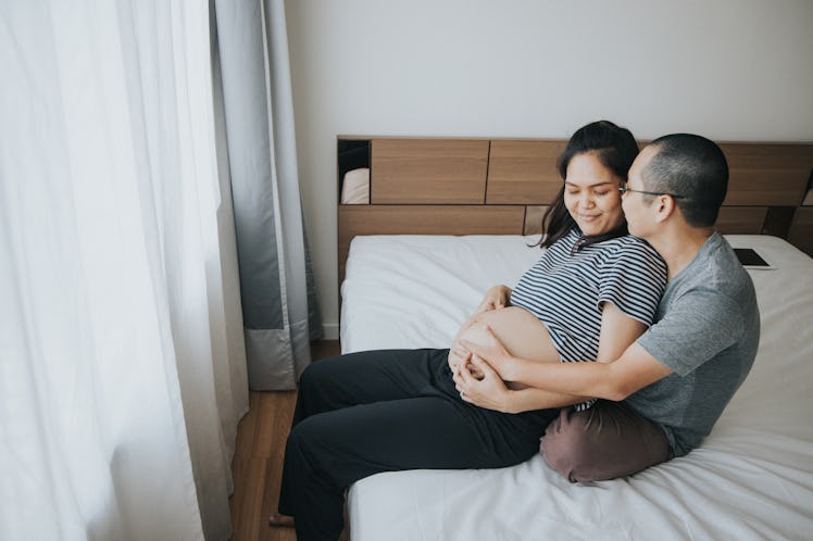 A man sits behind his pregnant wife on their bed, holding her.