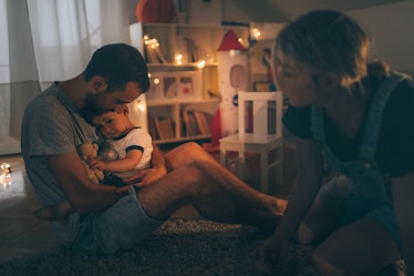 A father holds his sick child in his arms in the kid's bedroom as the mother sits with them.