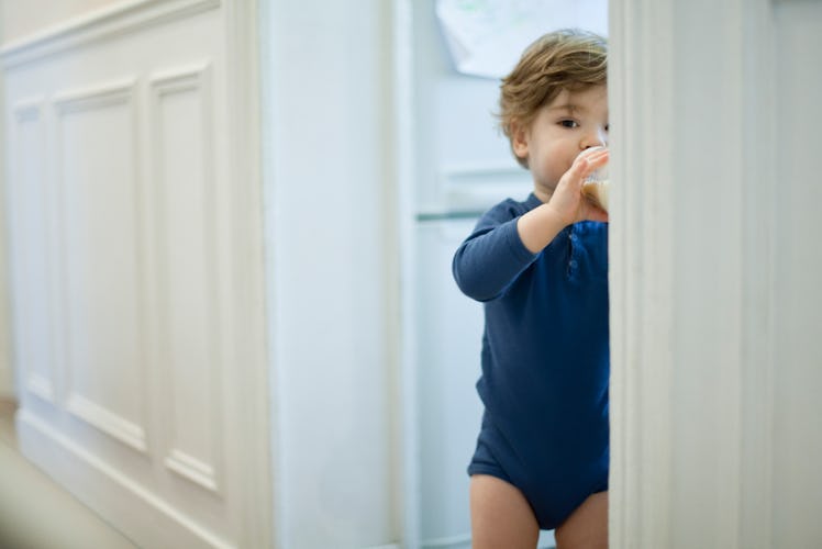 A toddler standing in a doorway while drinking milk from a bottle.