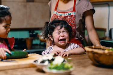 A toddler throwing a tantrum at the dinner table while their mother prepares food.
