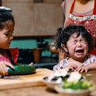 A toddler throwing a tantrum at the dinner table while their mother prepares food.