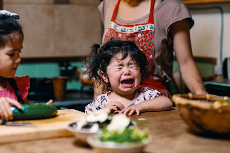 A toddler throwing a tantrum at the dinner table while their mother prepares food.