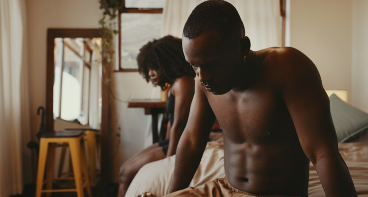 A shirtless man sits dejected at the food of the bed, with a woman sitting on the side of the bed.